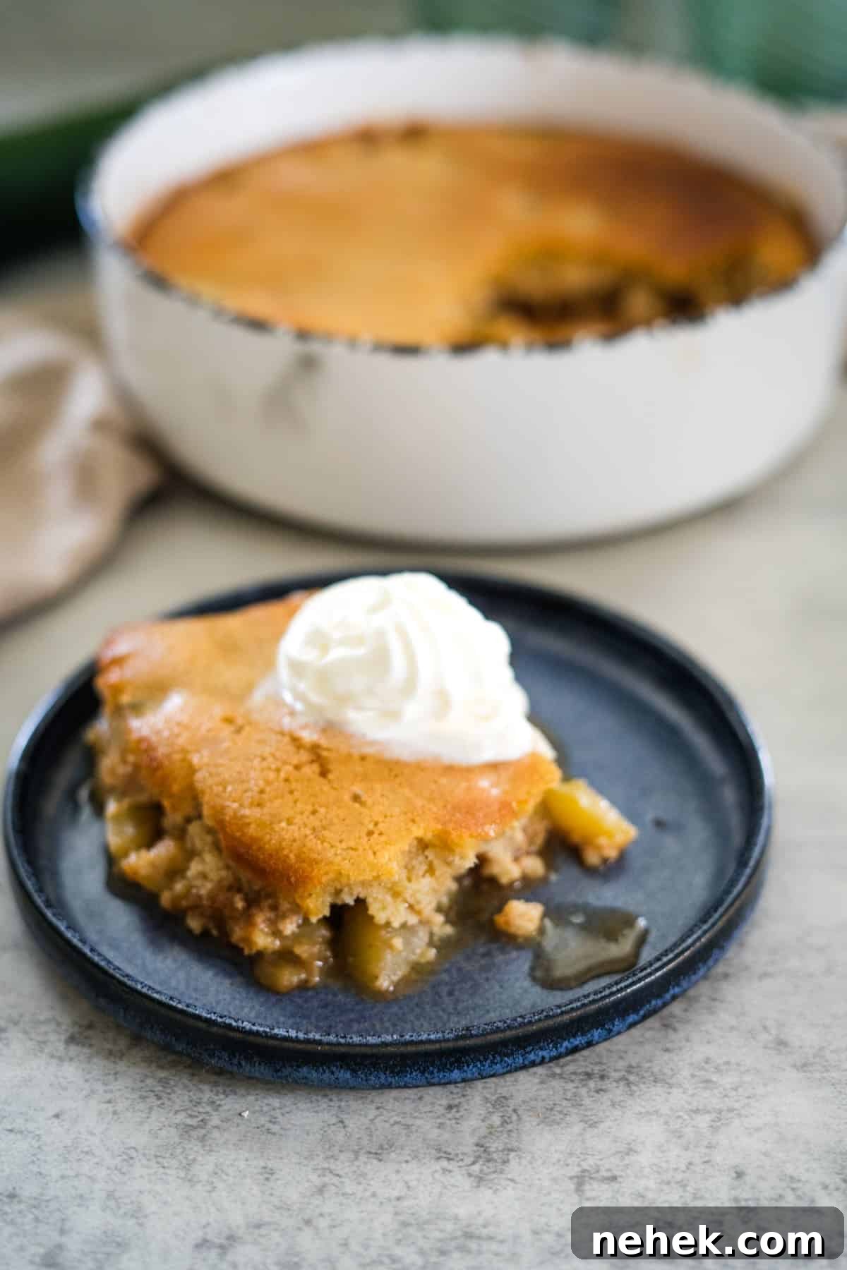 A slice of zucchini cobbler topped with whipped cream sits on a blue plate, with the remaining cobbler in a white dish in the background.
