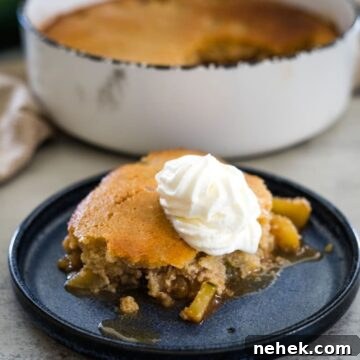 A slice of apple cobbler topped with whipped cream sits on a blue plate, with a larger dish of cobbler in the background.