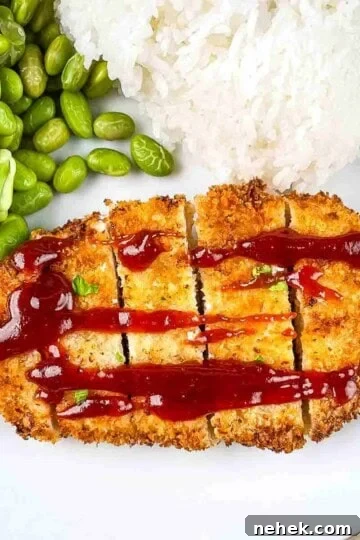 Golden crispy Chicken Katsu served on a white plate with a side of steamed white rice and green edamame, garnished with a lemon wedge and drizzled with tonkatsu sauce.