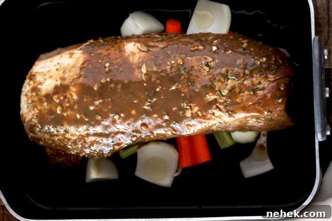 A raw pork loin roast sitting on top of carrots, onions and celery inside a roasting pan