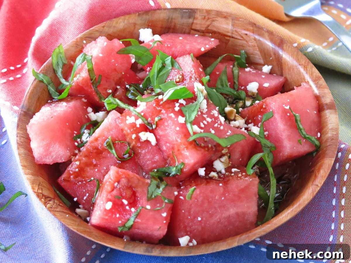 Chunks of watermelon topped with basil, balsamic glaze, and crumbled feta cheese in a brown bowl on a colorful napkin.