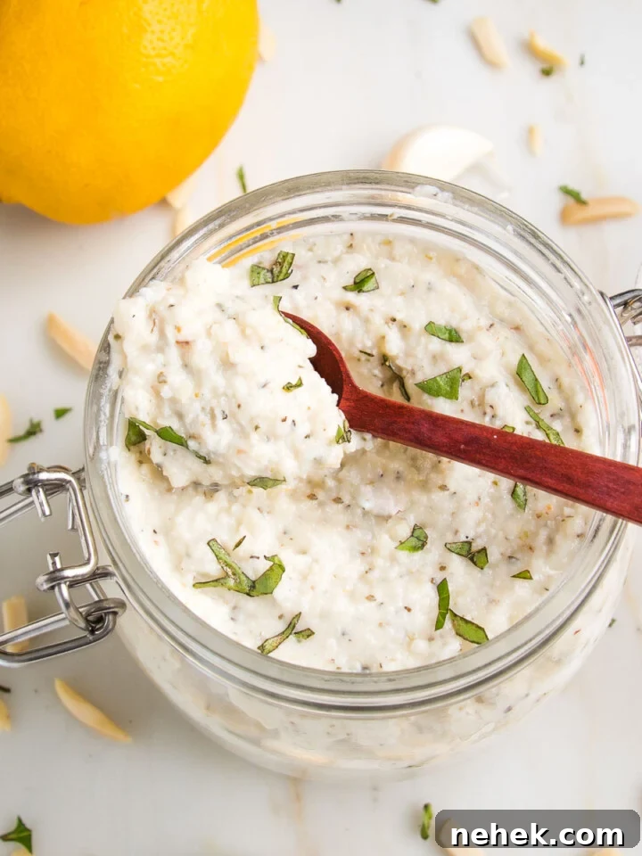 glass jar filled with vegan ricotta, sprinkled with fresh herbs...a spoon is scooping some out. THere are slivered almonds, garlic cloves and a lemon in the background.