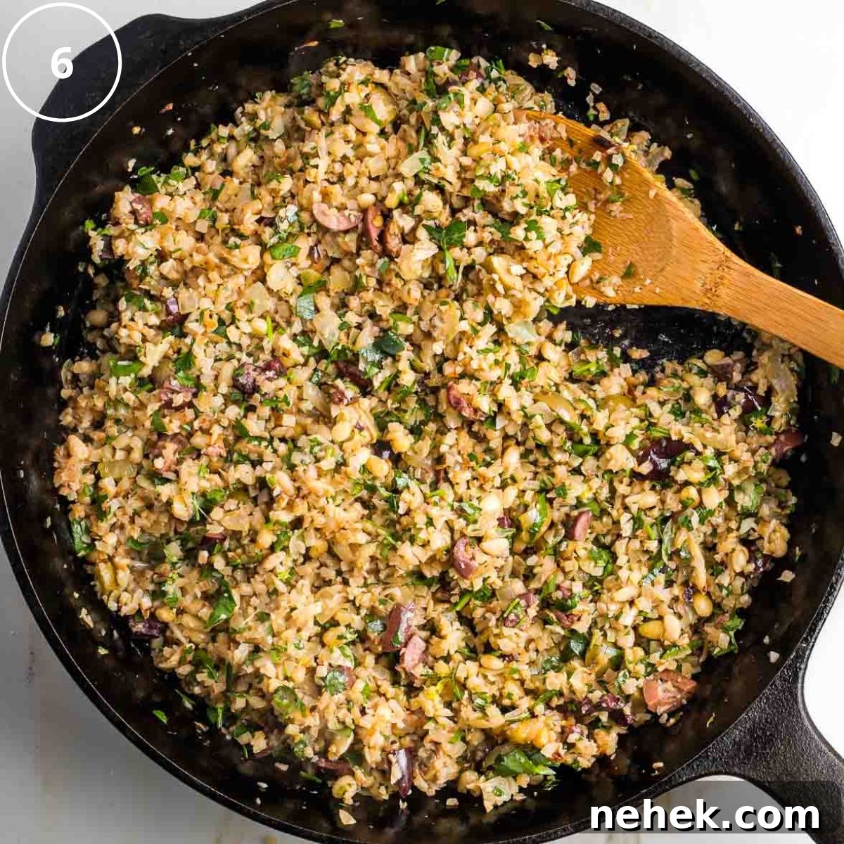 Mediterranean Cauliflower Rice Recipe with Sun‑Dried Tomatoes & Olives 9 Step 6: Wooden spoon stirring mediterranean cauliflower rice in a cast iron skillet with olives, fresh herbs and pine nuts.