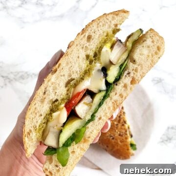 a hand holding up a sandwich with roasted vegetables, brie, and pesto