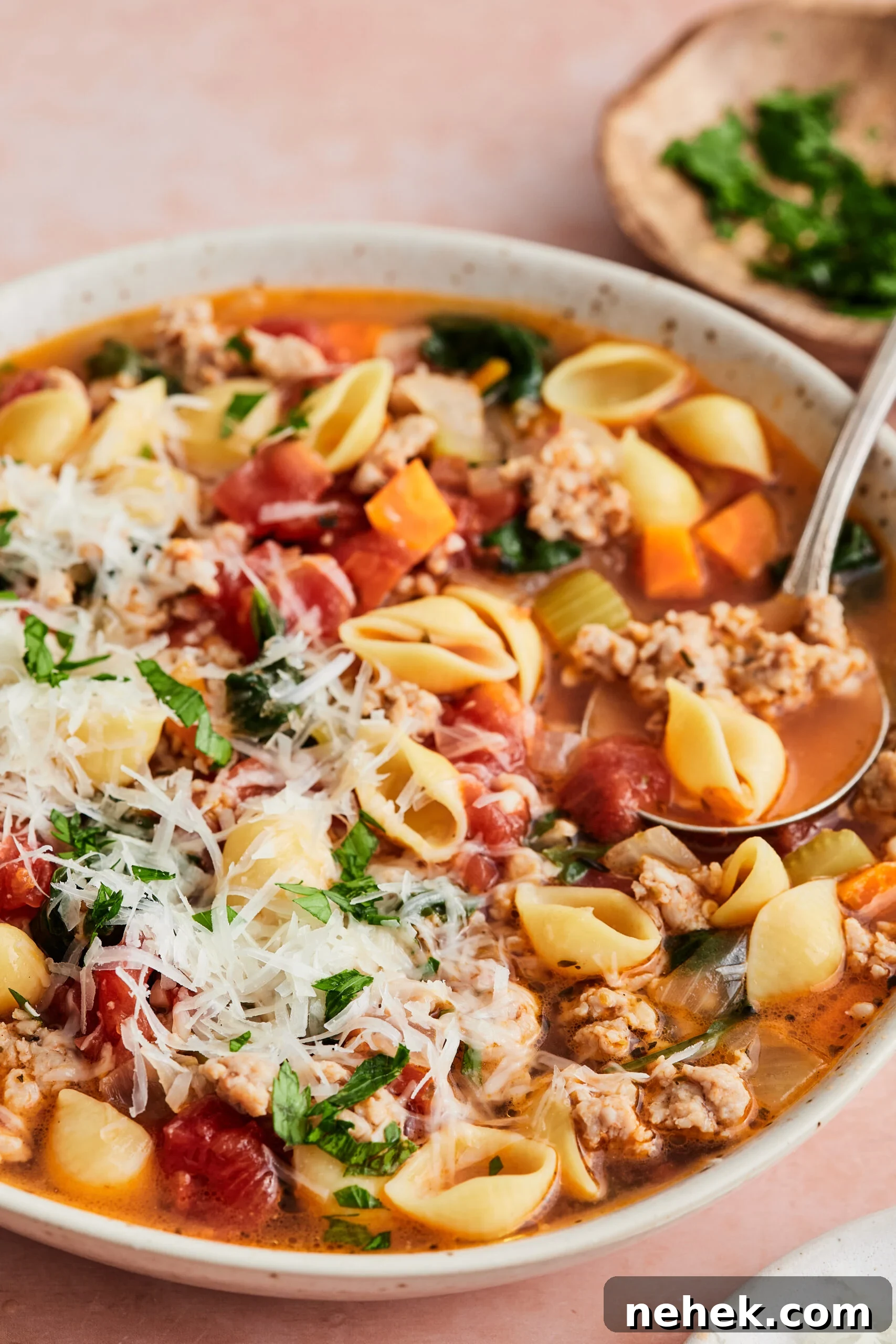 Bowl of Italian sausage soup topped with cheese and fresh herbs, with a spoon scooping out a serving against a light pink background.