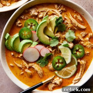 A bowl of keto white chicken chili with shredded chicken, avocado slices, jalapeños, cilantro, radish, and lime. Next to it is a glass of water with lime slices. Everything is set on a textured pinkish surface.