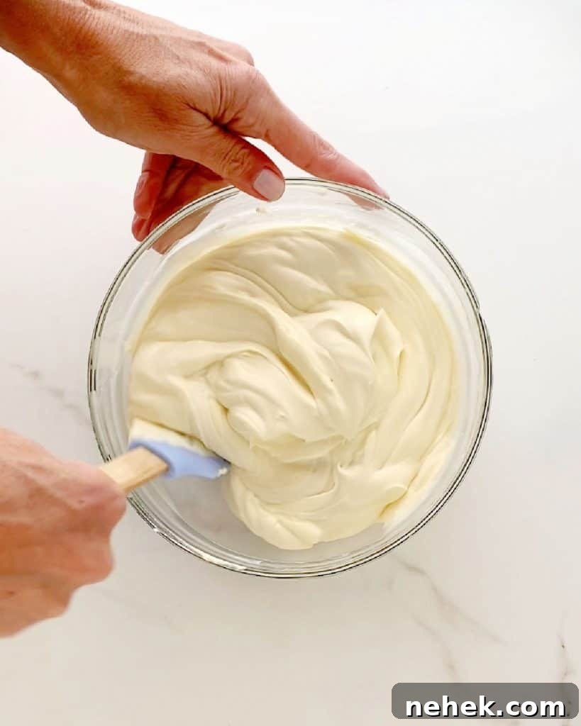 Mixing cheesecake ice cream mixture in a glass bowl with a spatula. White marbled surface.