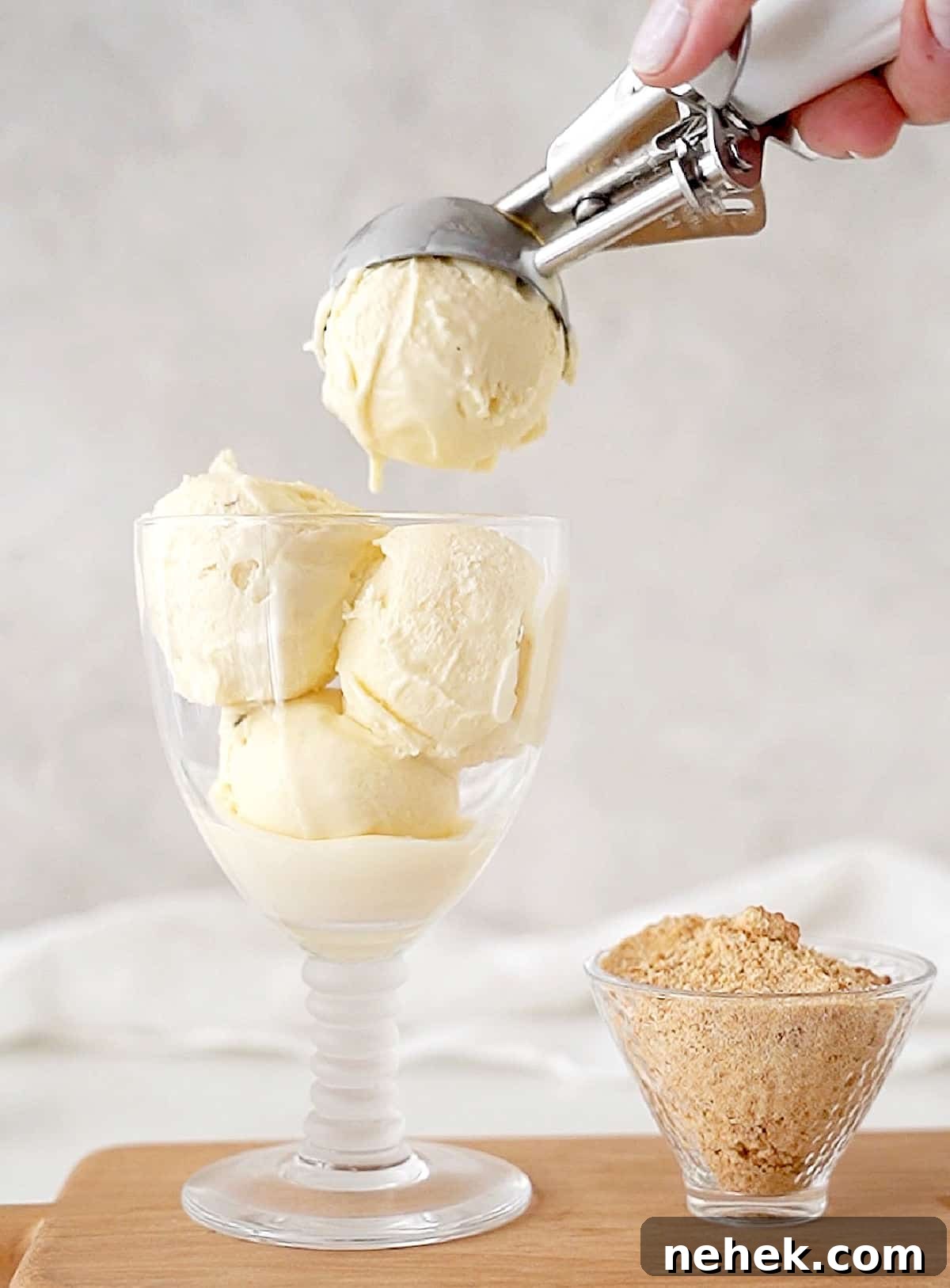 A glass with vanilla ice cream being scooped. Bowl with crumbs, wooden surface, grey background. 