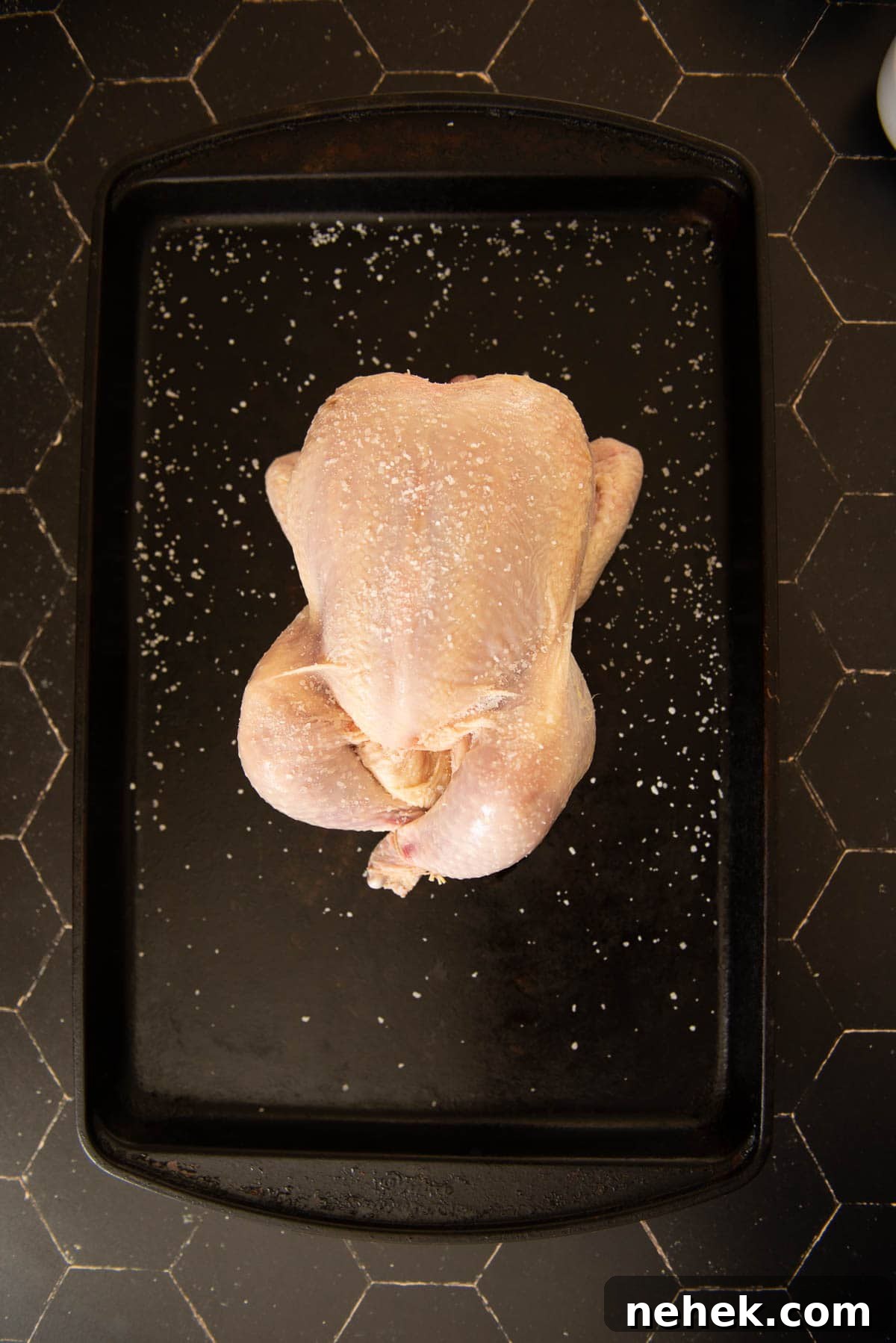 A chicken is sitting on a black baking tray, salted.