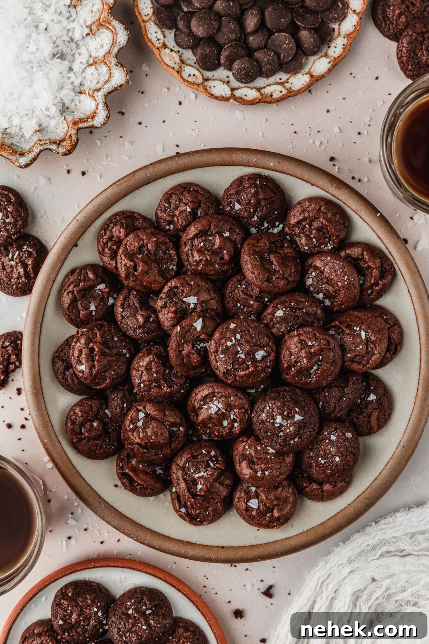 A tan plate of mini chocolate brownie crinkle cookies next to cups of coffee, a white linen, and brown bowls of cookies, chocolate, and salt on a tan counter.