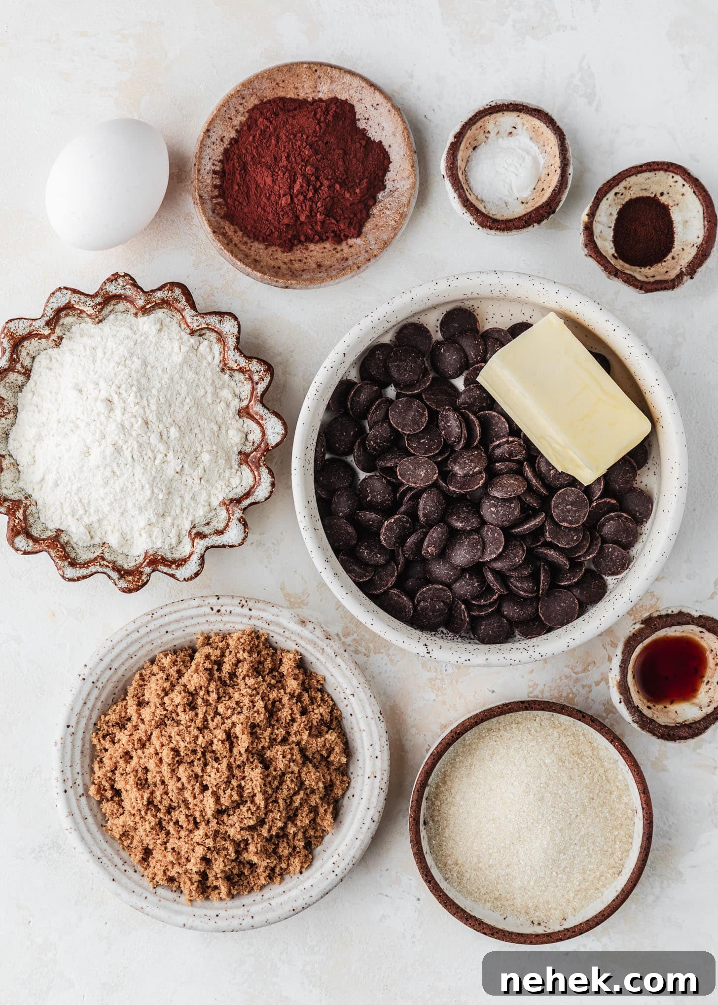 White and brown bowls of chocolate chips, butter, flour, cocoa, espresso powder, salt, baking soda, brown sugar, granulated sugar, and vanilla extract next to an egg on a tan table.