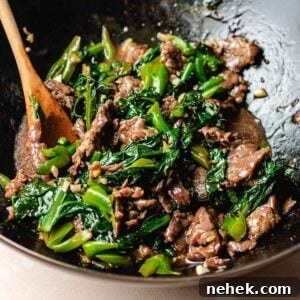 A close-up of Beef with Chinese broccoli stir-fry in a wok, featuring sliced beef, and leafy Chinese broccoli greens being mixed with a wooden spoon. The ingredients are coated in a glossy sauce.