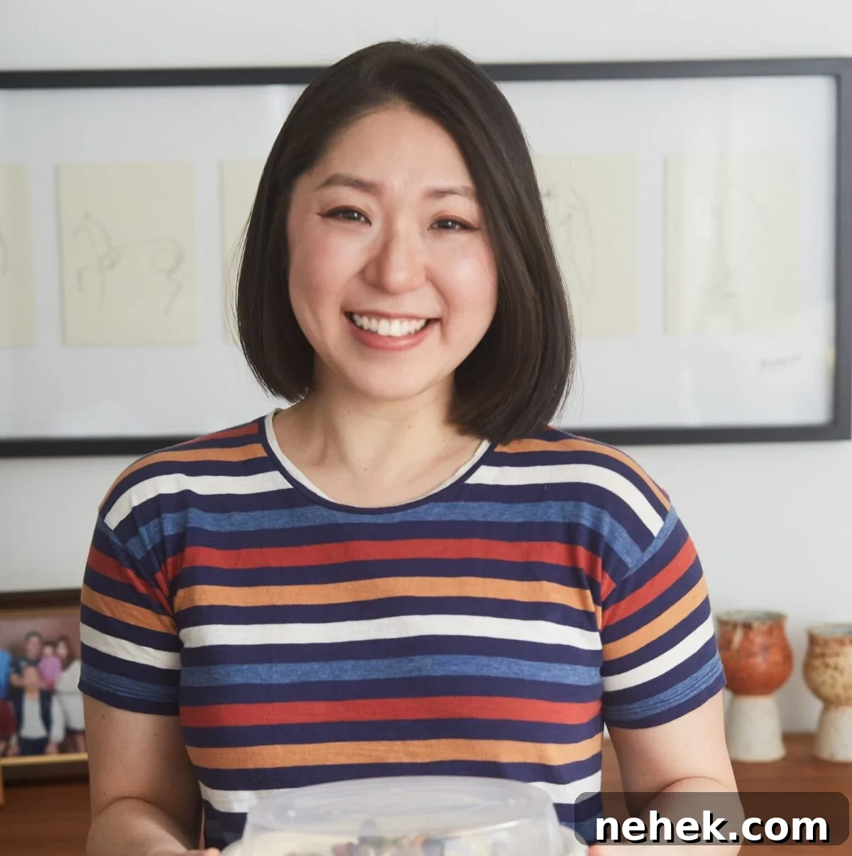ChihYu Smith with shoulder-length dark hair, wearing a striped shirt, smiles at the camera while holding a container of Creamy Roasted Sesame Dressing. Framed drawings and a family photo are visible on the wall and table behind her.