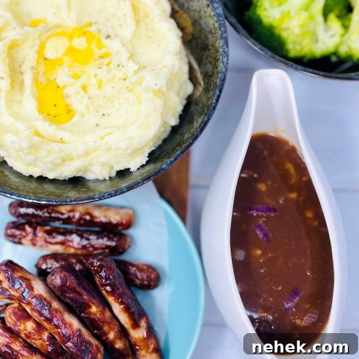 Image shows a Gravy Boat with red onion gravy inside, to the left there is a blue plate with several sausages on, above the plate there is a black bowl of mash, to the right of the mash there is a black bowl of brocolli