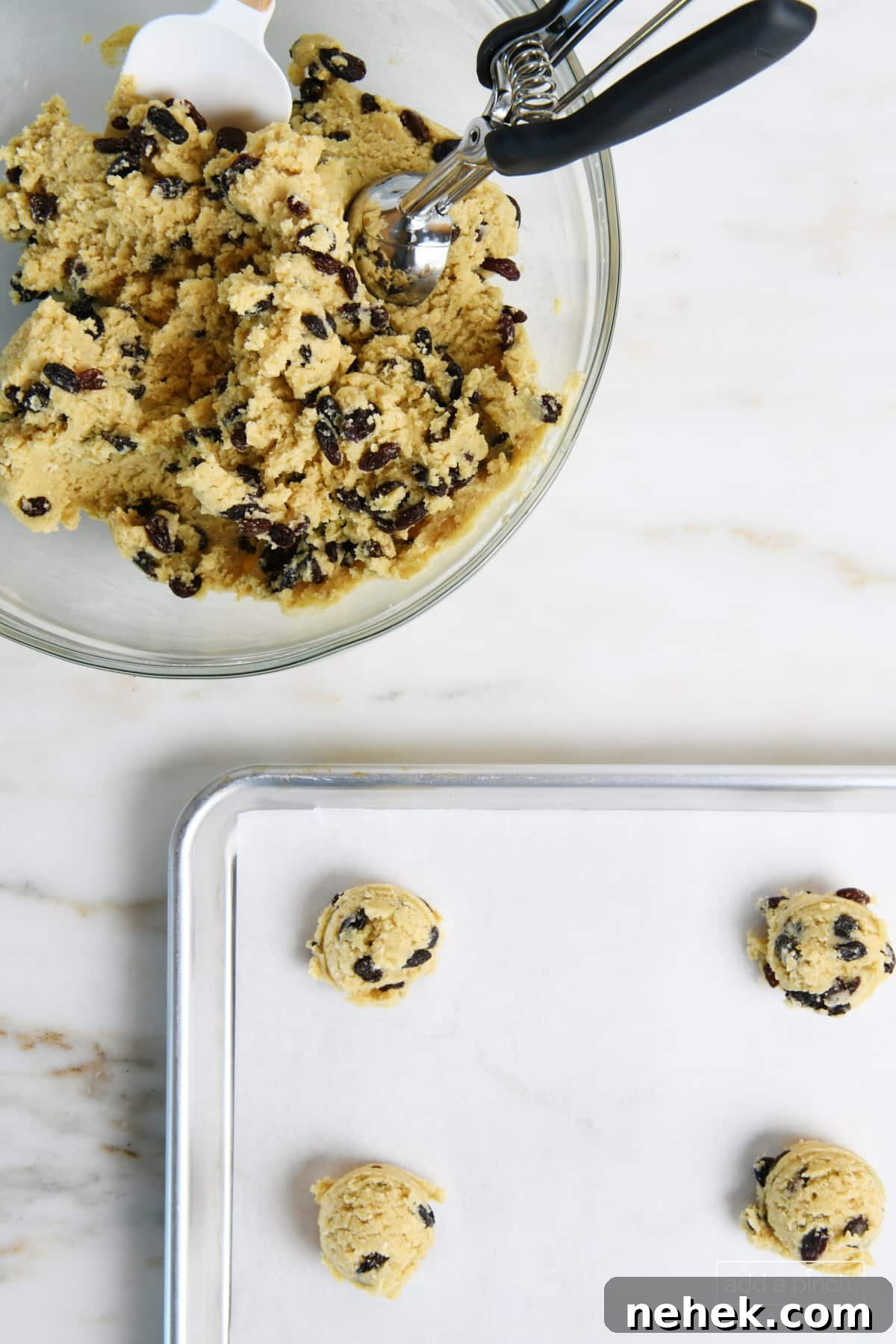 Oatmeal raisin cookie dough in glass bowl with scoop next to a parchment lined baking sheet with small scoops of cookie dough balls placed on it.