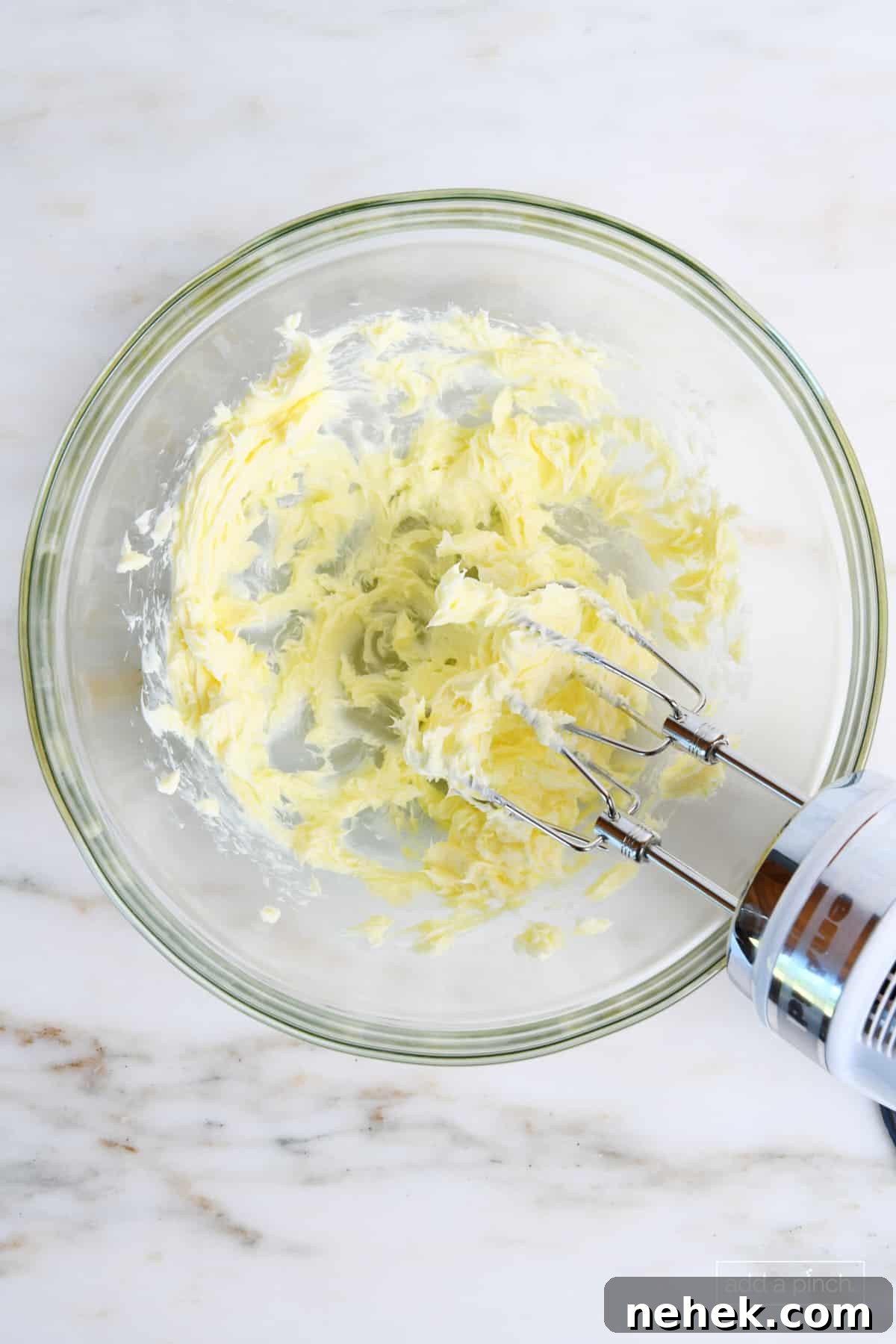 Softened butter is being creamed with a hand mixer in a glass bowl.