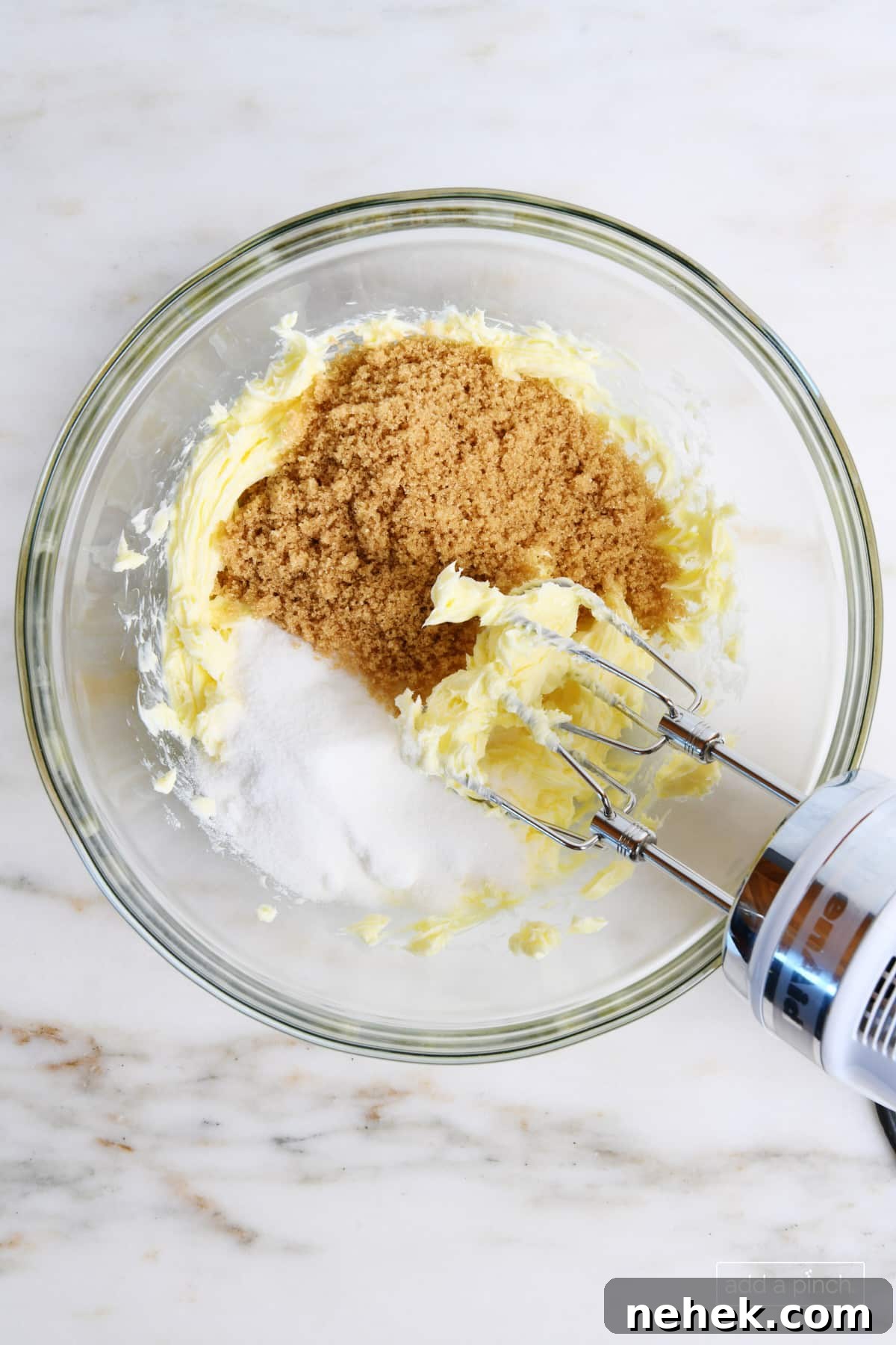 Glass mixing bowl holds softened butter, brown sugar and granulated sugar being creamed together with a hand mixer.
