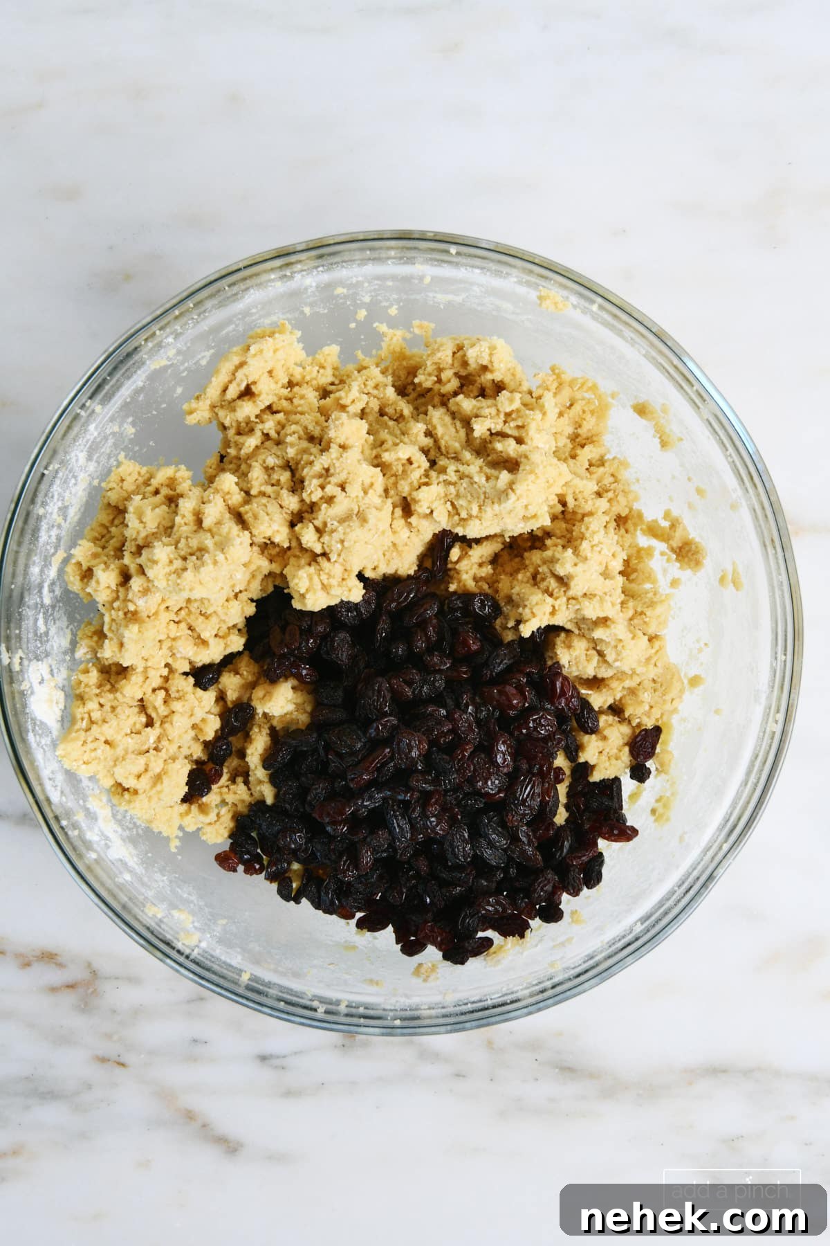 Glass bowl of cookie dough has raisins added ready to mix in to make oatmeal raisin cookie dough.