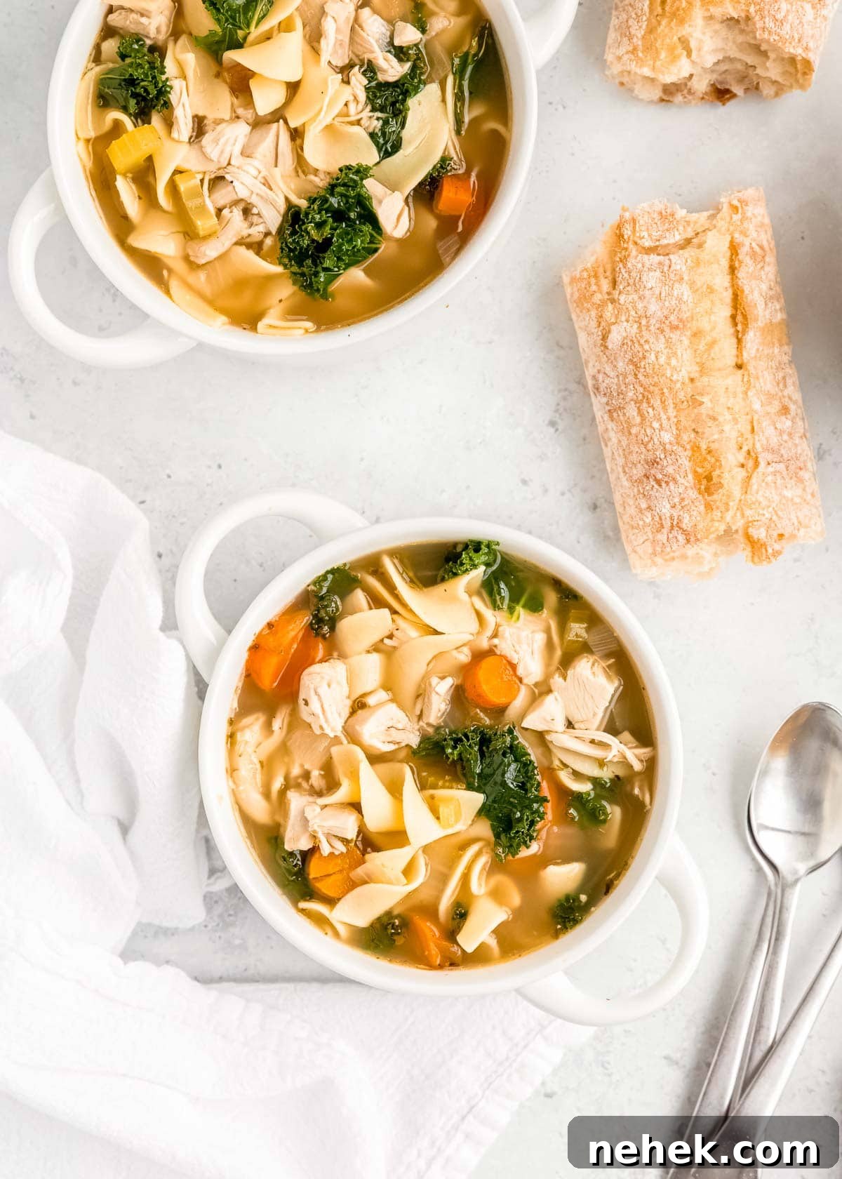 Classic Homemade Chicken Noodle Soup Recipe for Comforting Meals 11 two bowls of chicken noodle soup with egg noodles and kale with white napkins, silver spoons, and two pieces of bread on a white table