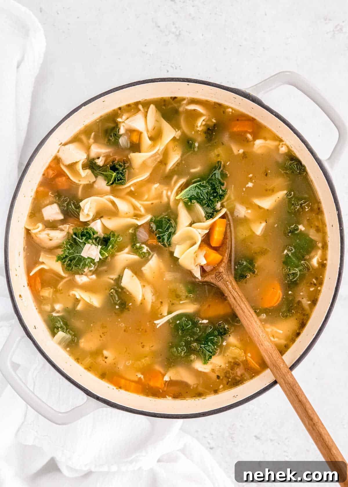 Classic Homemade Chicken Noodle Soup Recipe for Comforting Meals 13 overhead shot of a large pot of chicken noodle soup with a wooden spoon picking up egg noodles, shredded chicken, carrots, and kale in a delicious broth.