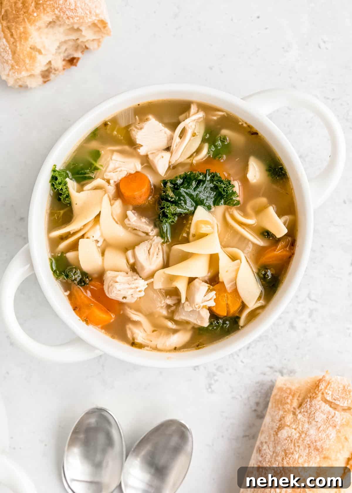 Classic Homemade Chicken Noodle Soup Recipe for Comforting Meals 14 overhead shot of a white bowl filled with classic chicken noodle soup with egg noodles. bread and spoons shown in the background