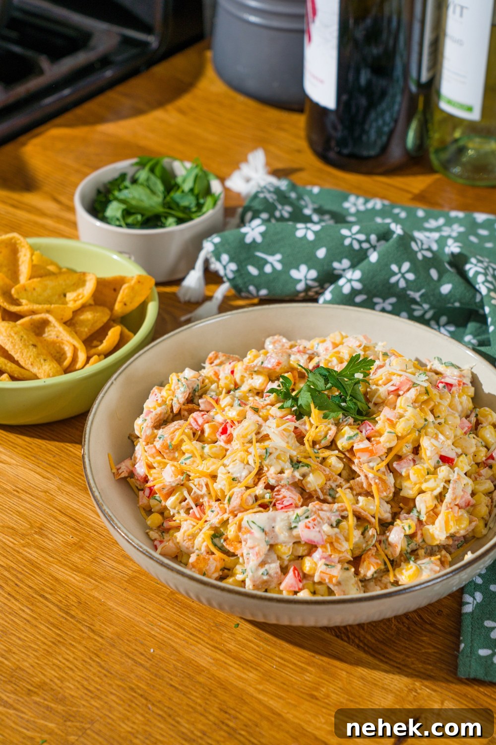 Spicy Corn Salad with Chili Cheese Fritos Crunch 2 A white bowl with corn salad with chili cheese fritos garnished with fresh parsley. The bowl is on a wooden counter with a green floral napkin and a bowl of garnishes.