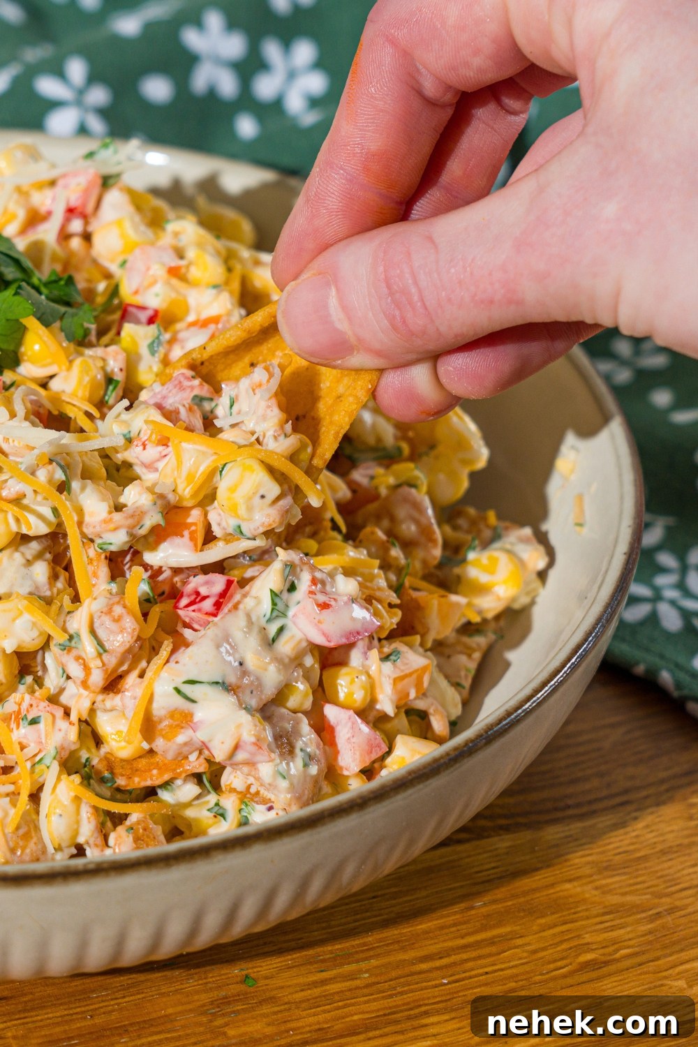 Spicy Corn Salad with Chili Cheese Fritos Crunch 11 A white bowl with corn salad with chili cheese fritos garnished with fresh parsley. A hand is dipping a chip into the bowl. The bowl is on a wooden counter with a green floral napkin.