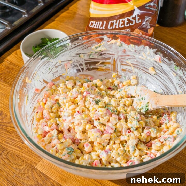 A glass bowl with a wooden spoon mixing corn, shredded cheese, and diced peppers in a sour cream dressing. The bowl is on a wooden counter with a bag of fritos.