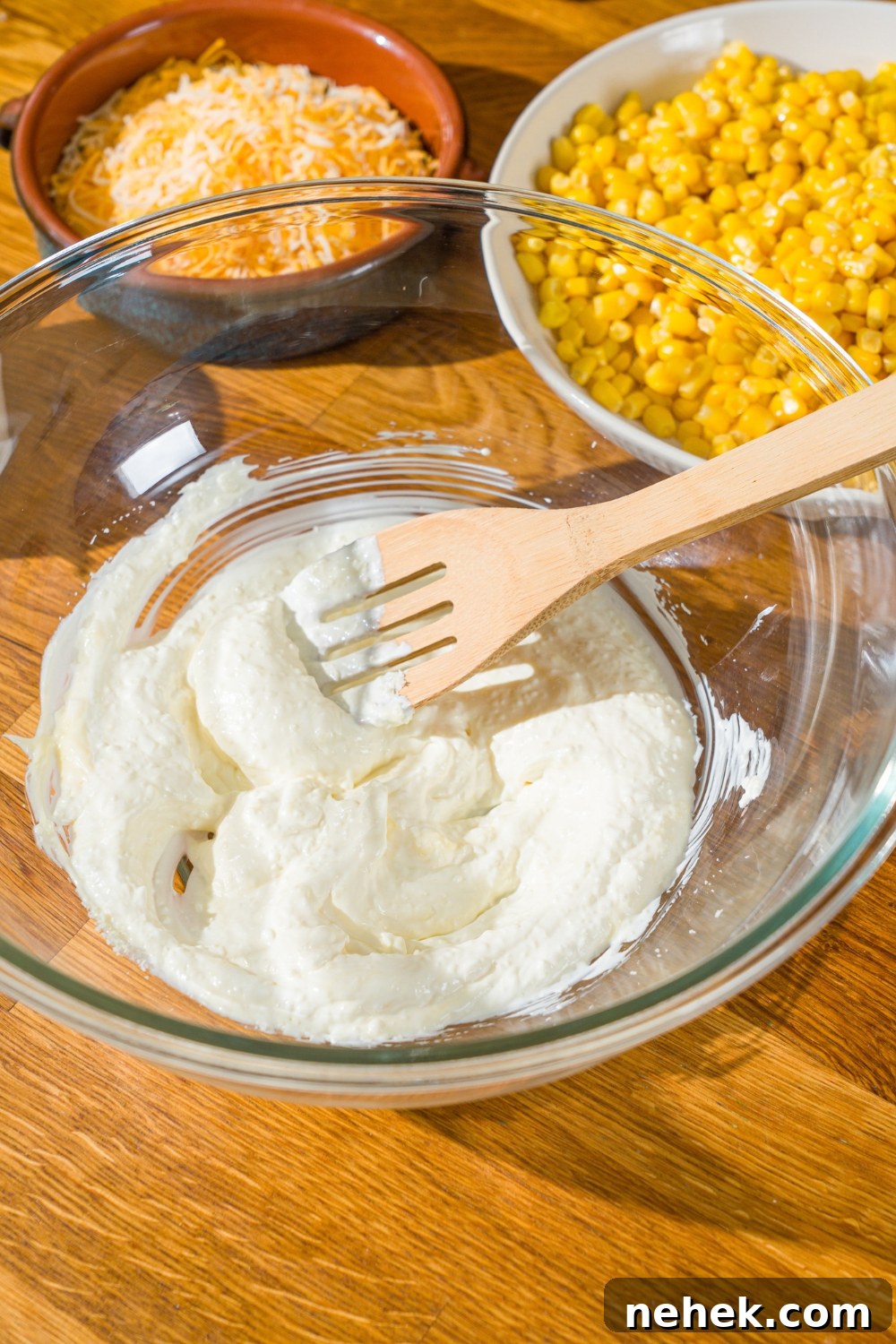 Spicy Corn Salad with Chili Cheese Fritos Crunch 5 A glass bowl with a wooden spoon mixing corn salad sour cream dressing. The bowl is on a wooden counter with bowls of ingredients.