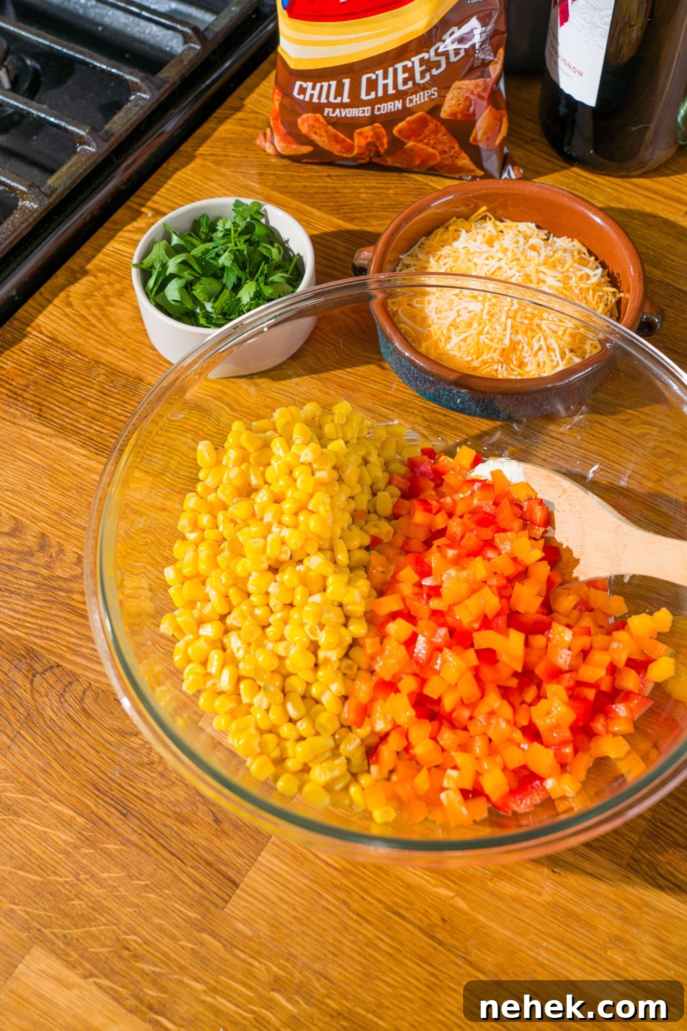 Spicy Corn Salad with Chili Cheese Fritos Crunch 6 A glass bowl with a spoon mixing corn and diced red and yellow bell pepper. The bowl is on a wooden counter with bowls of ingredients.