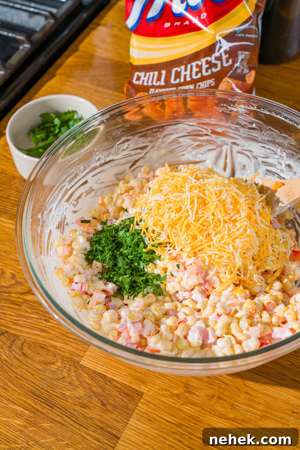 Spicy Corn Salad with Chili Cheese Fritos Crunch 7 A glass bowl with corn and diced peppers tossed in a sour cream dressing. There is shredded cheese and chopped parsley on top. The bowl is on a wooden counter.