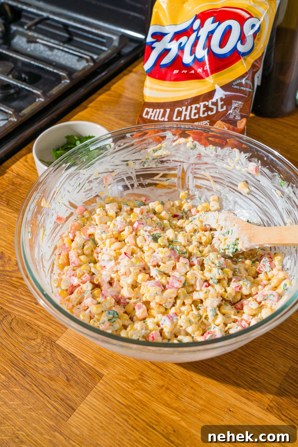 Spicy Corn Salad with Chili Cheese Fritos Crunch 8 A glass bowl with a wooden spoon mixing corn, shredded cheese, and diced peppers in a sour cream dressing. The bowl is on a wooden counter with a bag of fritos.