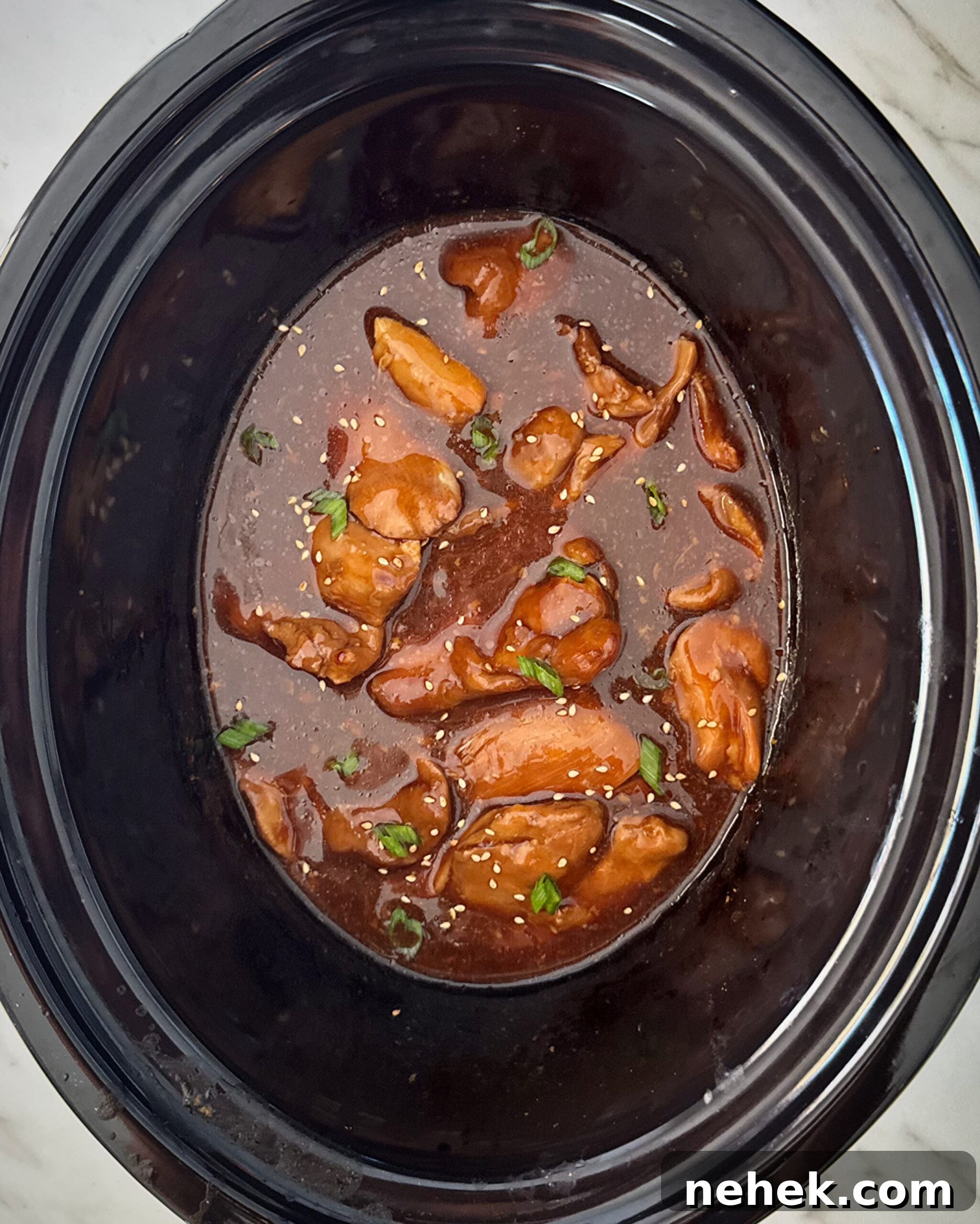 overhead shot of cooked teriyaki chicken in a black slow cooker garnished with green onion and sesame seeds