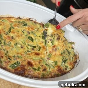 A person with red nail polish slices a piece of zucchini and cheese casserole from a white baking dish. The scene, perfect for serving the BEST Vegetable Frittata, features fresh greens in the background on a wooden countertop.