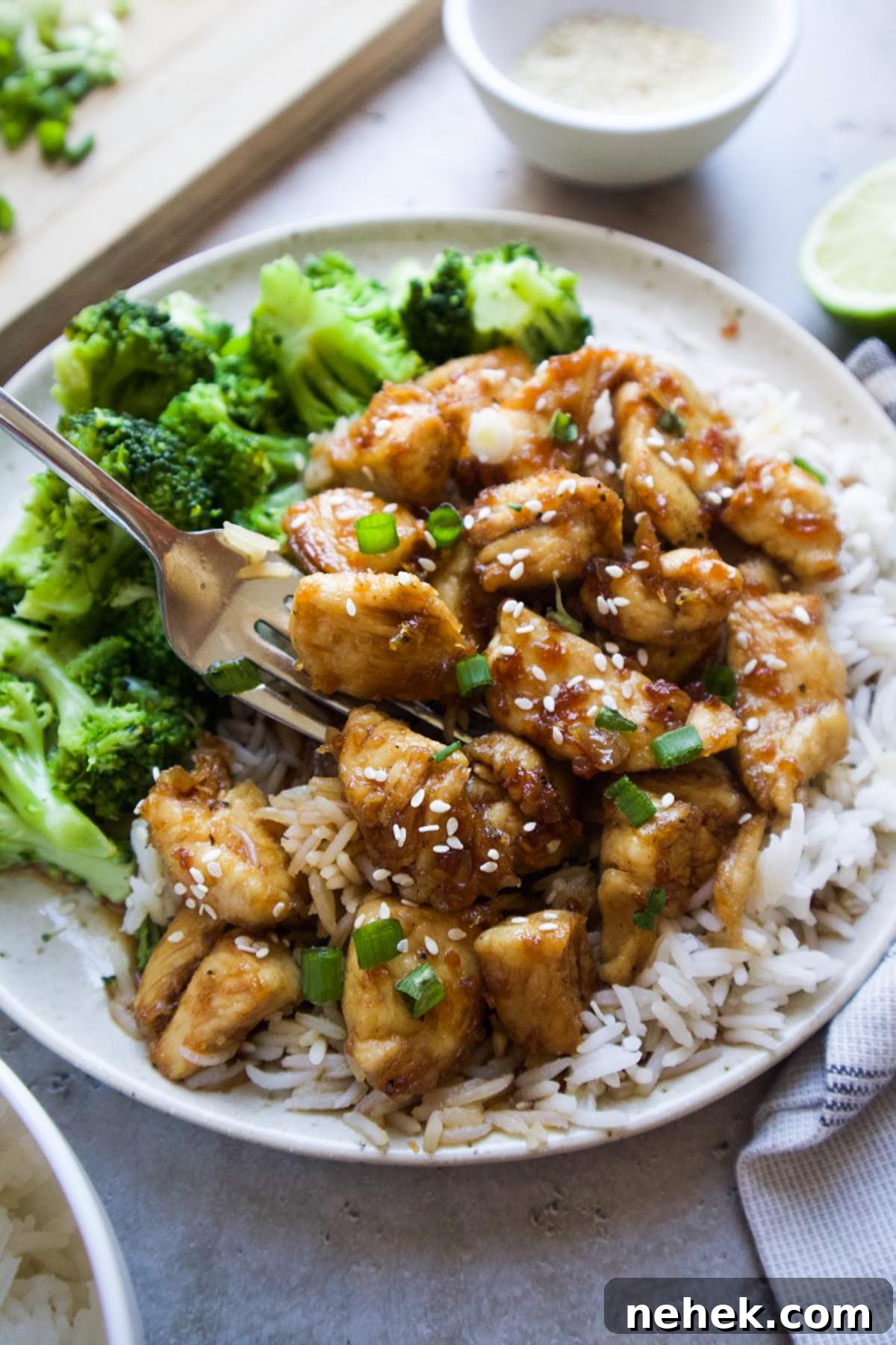 white plate with chicken over top of white rice and a side of broccoli topped with sesame seeds and green onion.