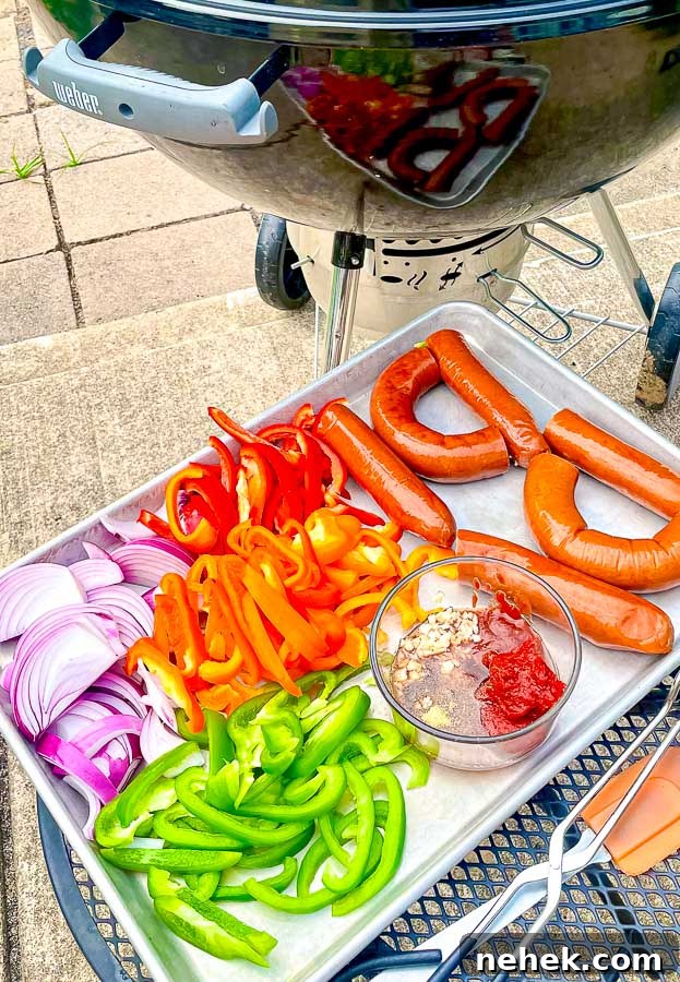 ingredients for grilled sausage with onions and peppers next to a weber grill