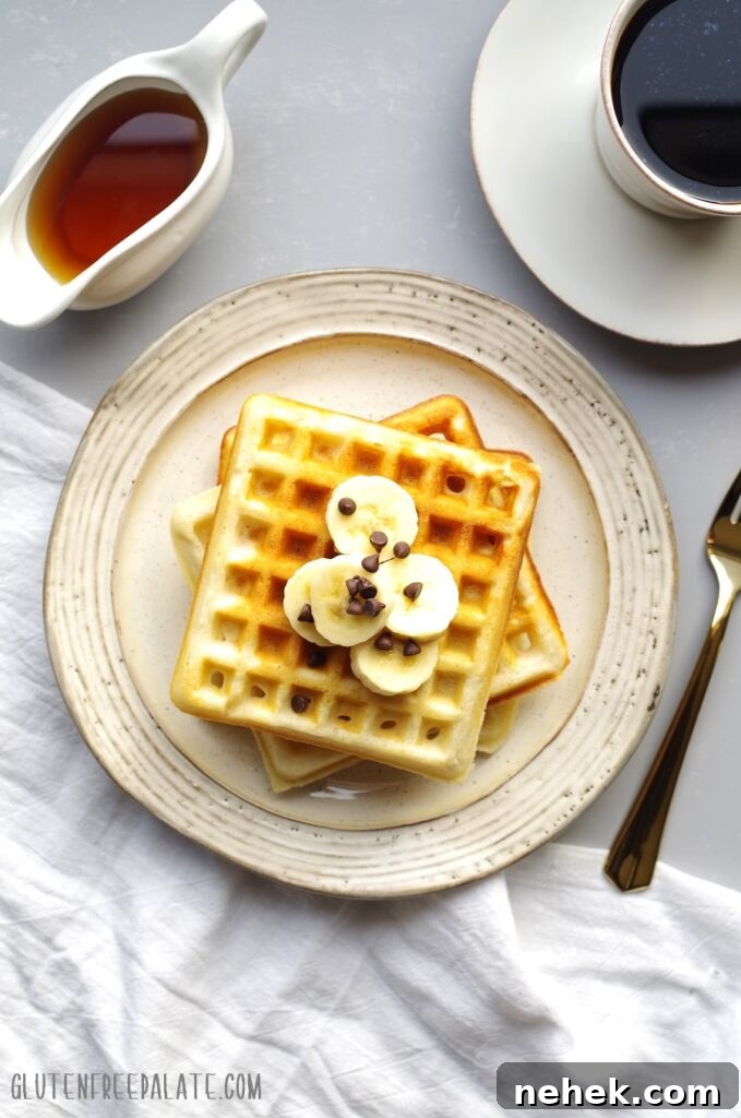 Three gluten-free waffles stacked on a cream colored plate, topped with sliced bananas, next to a cup of coffee and a container of maple syrup.