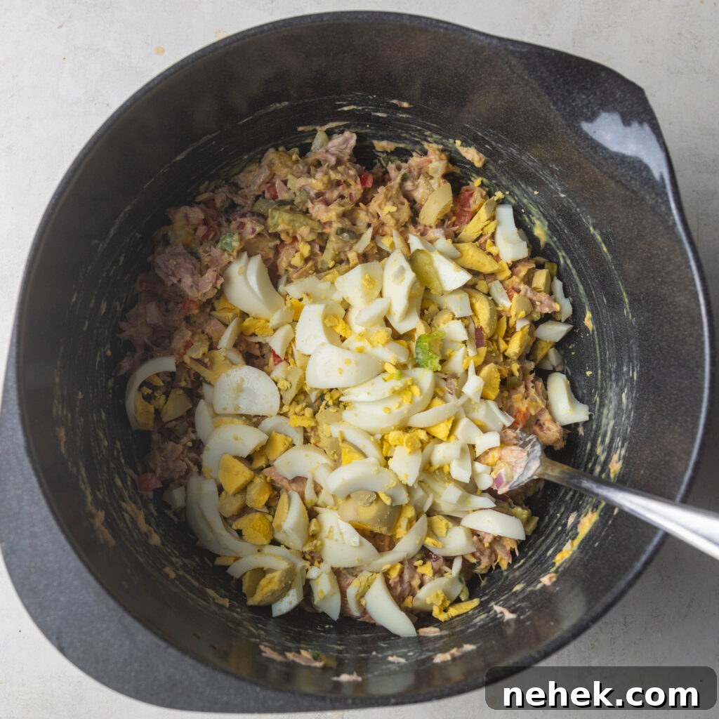 Adding mustard, mayonnaise, and chopped boiled eggs into bowl with tuna fish and diced vegetables. 