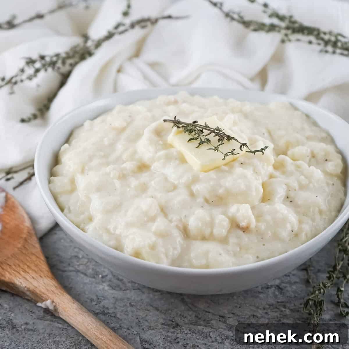 Creamy Mashed Potatoes and Parsnips Recipe 6 Close up shot of parsnip and potato mash with a pad of butter and herbs in a white bowl with a wooden spoon on the side.