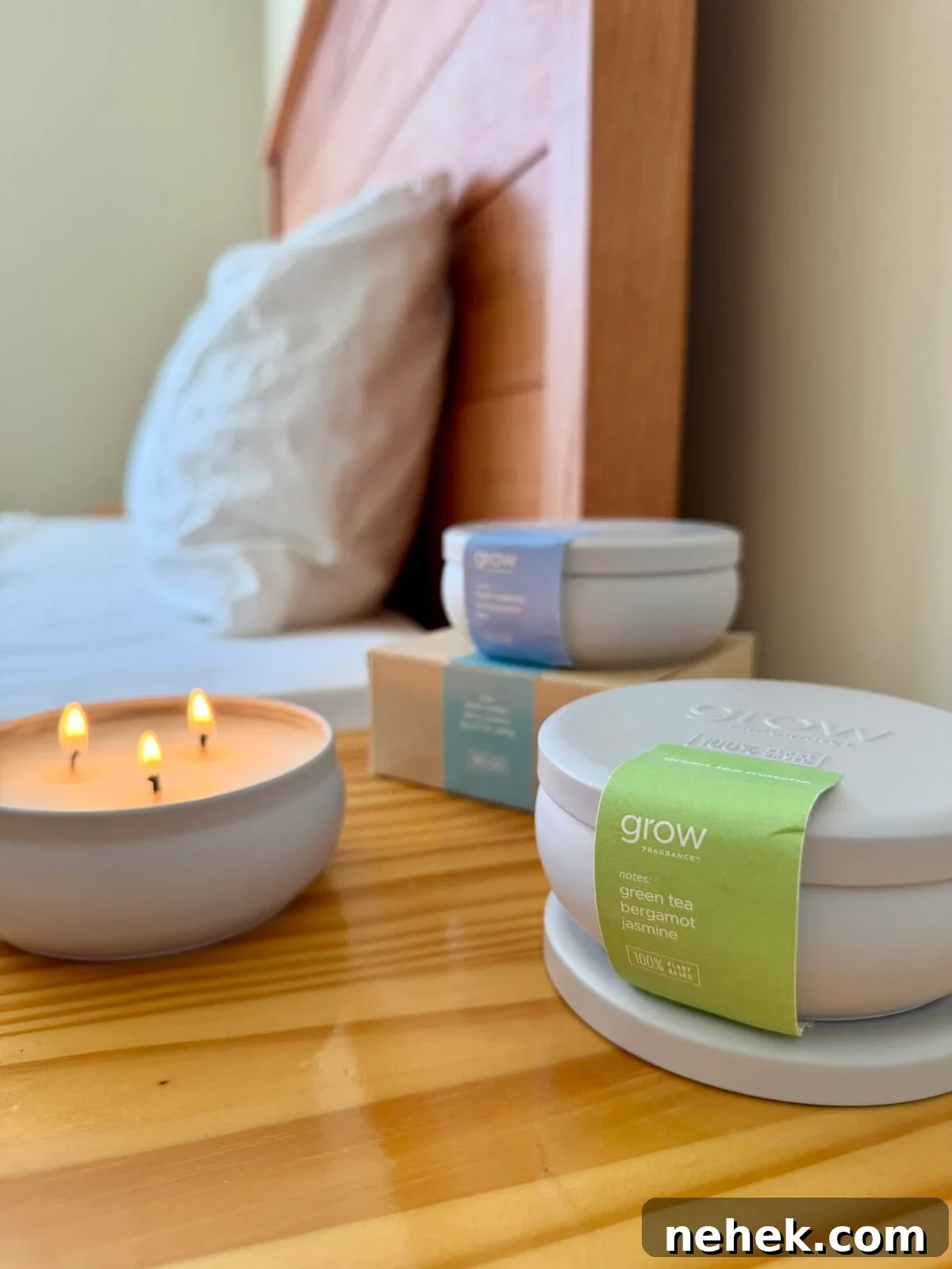 5 Non-Toxic Soy Candle Brands for Natural, Clean Fragrance 3 A lit three-wick candle sits on a wooden bedside table next to a bed with white pillows. Nearby are two closed round candle containers and a small cardboard box, all labeled grow with different scents.